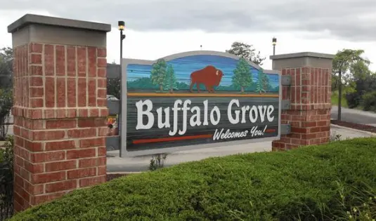 A large entrance sign reading Buffalo Grove Welcomes You with a graphic of a buffalo, flanked by two red brick pillars and green bushes.