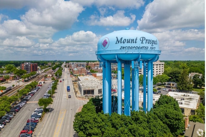 Aerial view of Mount Prospect, IL featuring the iconic blue water tower inscribed with 'Mount Prospect — Incorporated 1917,' surrounded by tree-lined streets, downtown buildings, and a partly cloudy sky